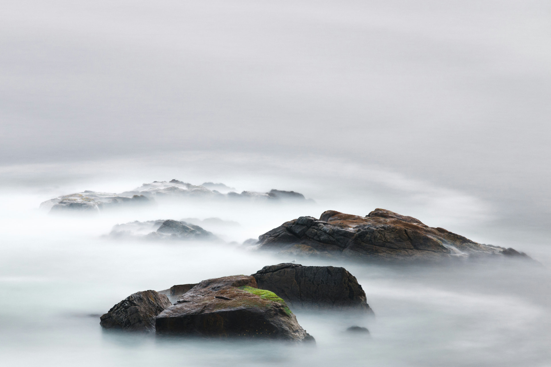 Dark craggy rocks emerging from soft white mist over a still sea
