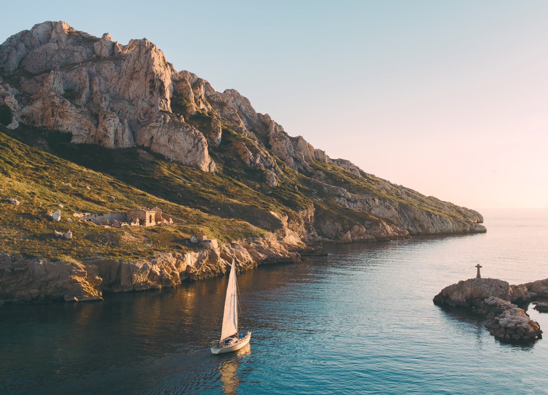 Sailboat gliding through calm coastal water at sunset beneath rocky cliffs